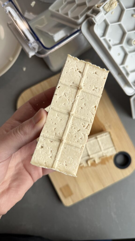 Selbstgemachter Tofublock in der Hand, im Hintergrund Tofu-Presse auf Holzbrett